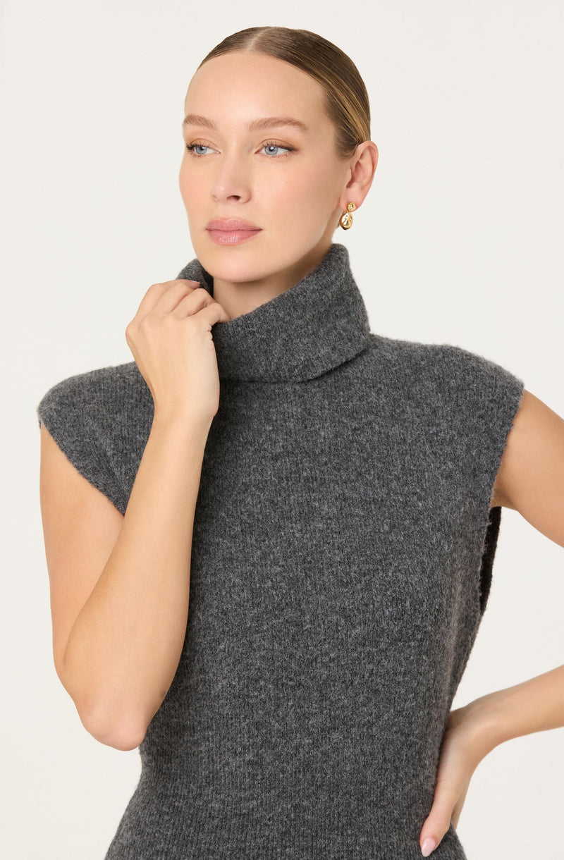 A woman is wearing a sleeveless, high-neck, dark gray knit dress with a ribbed turtleneck collar. She has gold hoop earrings and has her hair slicked back.