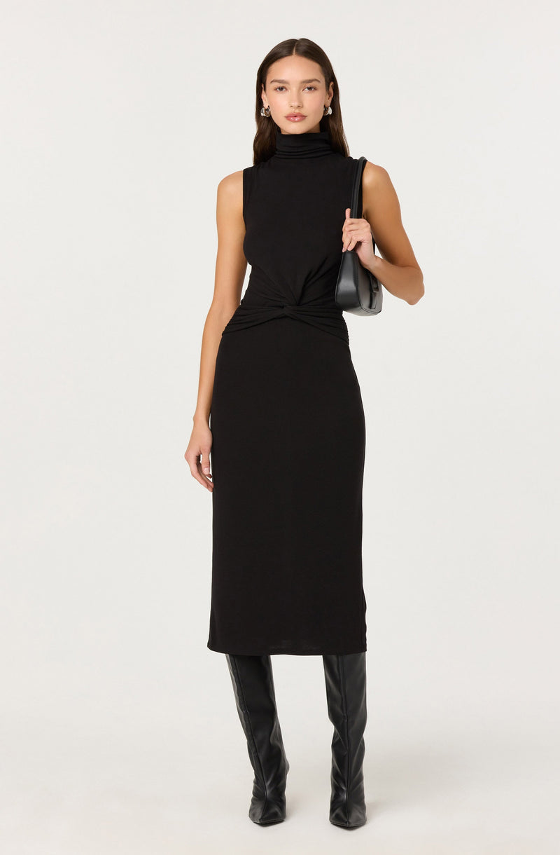A woman is wearing a sleeveless black turtleneck top paired with a high-waisted, ankle-length black skirt that has a gathered detail at the waist. She is also wearing black knee-high boots made of leather or a similar material. She carries a black handbag over her shoulder and is accessorized with earrings.