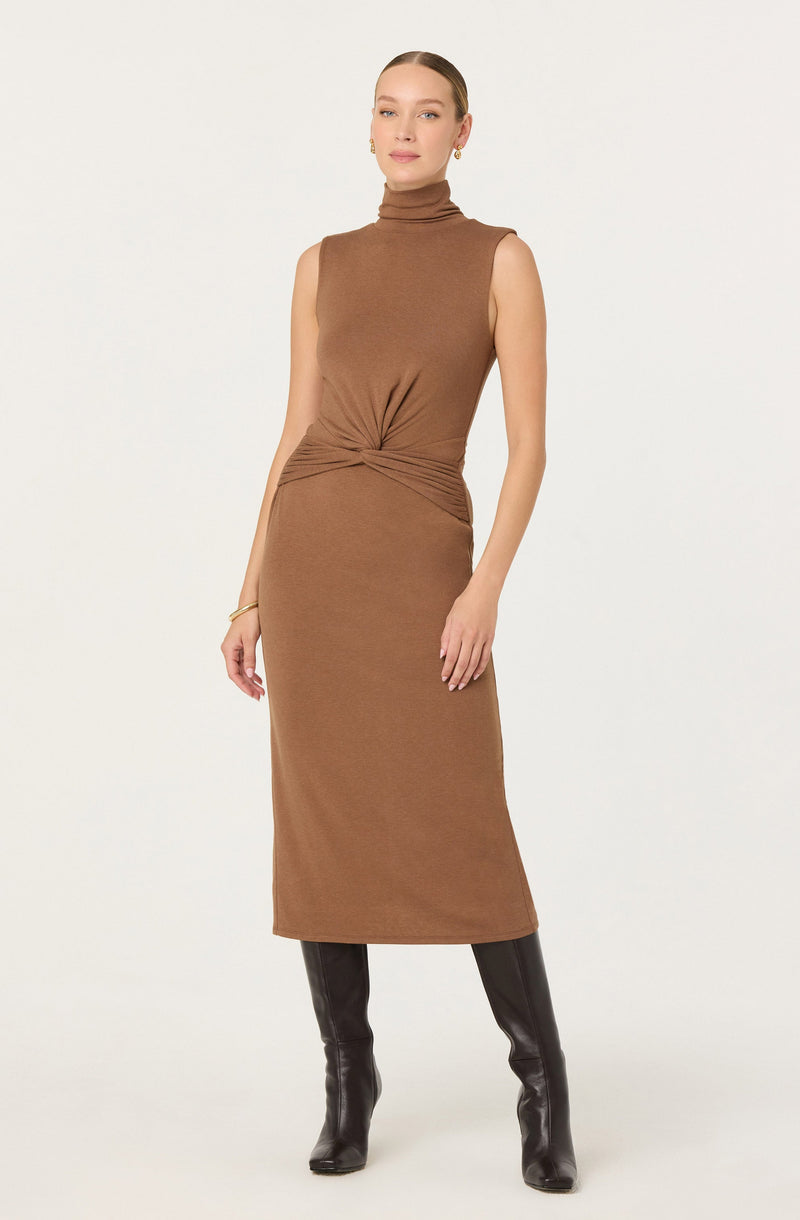 A woman is wearing a sleeveless, high-neck, camel-colored knit dress that extends to her mid-calf. The dress has a twisted knot detail at the waist, creating slight gathers on each side. She pairs the dress with tall, black leather knee-high boots.