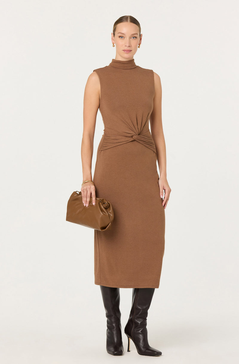 A woman is wearing a sleeveless, high-neck, brown knit dress that is fitted at the waist with a twisted knot detail. She accessorizes with small gold earrings, a gold bracelet, and carries a matching brown handbag. She is also wearing tall, black leather high-heeled boots.