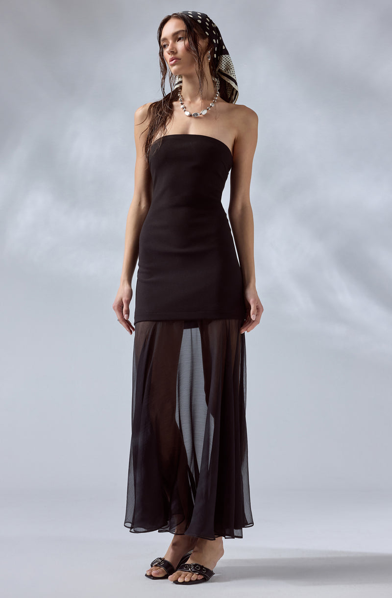 A woman is wearing a strapless black dress that is fitted at the top and extends down to her mid-thigh. She has on sheer, black, wide-leg pants that are see-through and flow to the floor. She accessorizes with a black and white patterned headscarf, a shell necklace, and black sandals with embellishments.