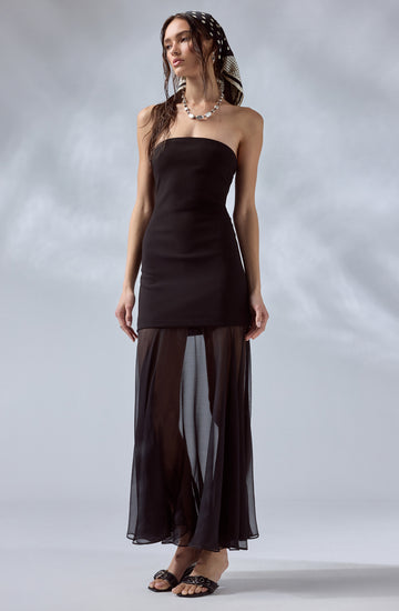 A woman is wearing a strapless black dress that is fitted at the top and extends down to her mid-thigh. She has on sheer, black, wide-leg pants that are see-through and flow to the floor. She accessorizes with a black and white patterned headscarf, a shell necklace, and black sandals with embellishments.