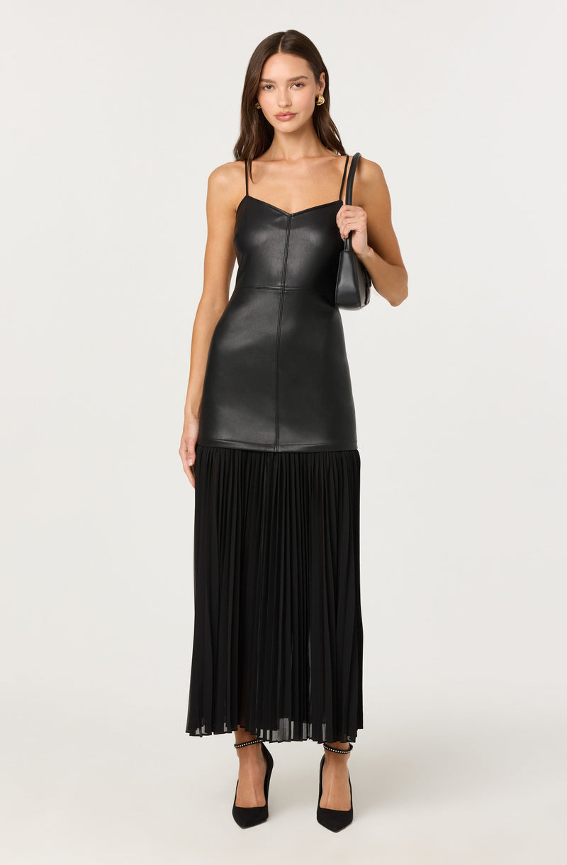 A woman is wearing a black spaghetti strap dress. The upper part of the dress appears to be made of a shiny leather-like material, while the lower part is a pleated fabric that reaches her ankles. She is also holding a small black shoulder bag and wearing black high heels.