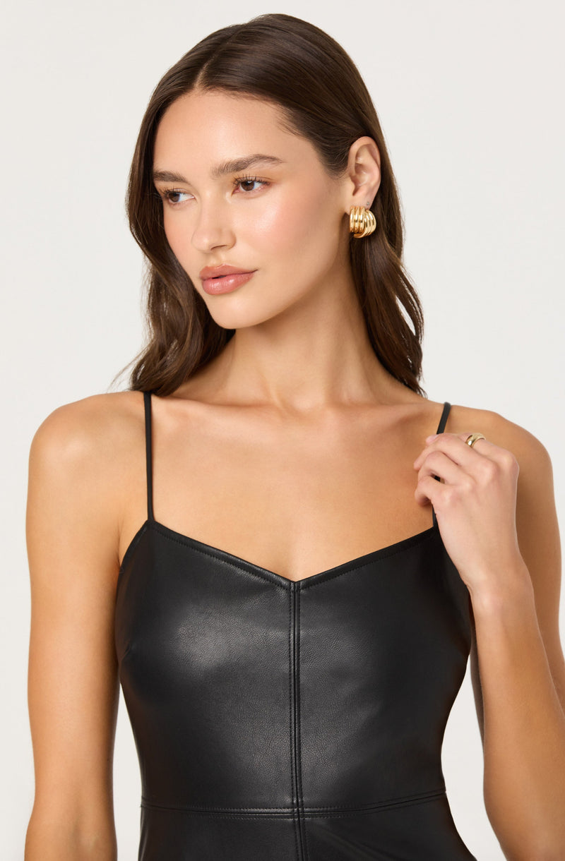 A woman is wearing a sleek, black, faux leather slip dress with thin spaghetti straps. She accessorizes with large, gold, statement earrings and a gold ring on her right hand. Her hair is styled in loose waves, and she has a natural makeup look.