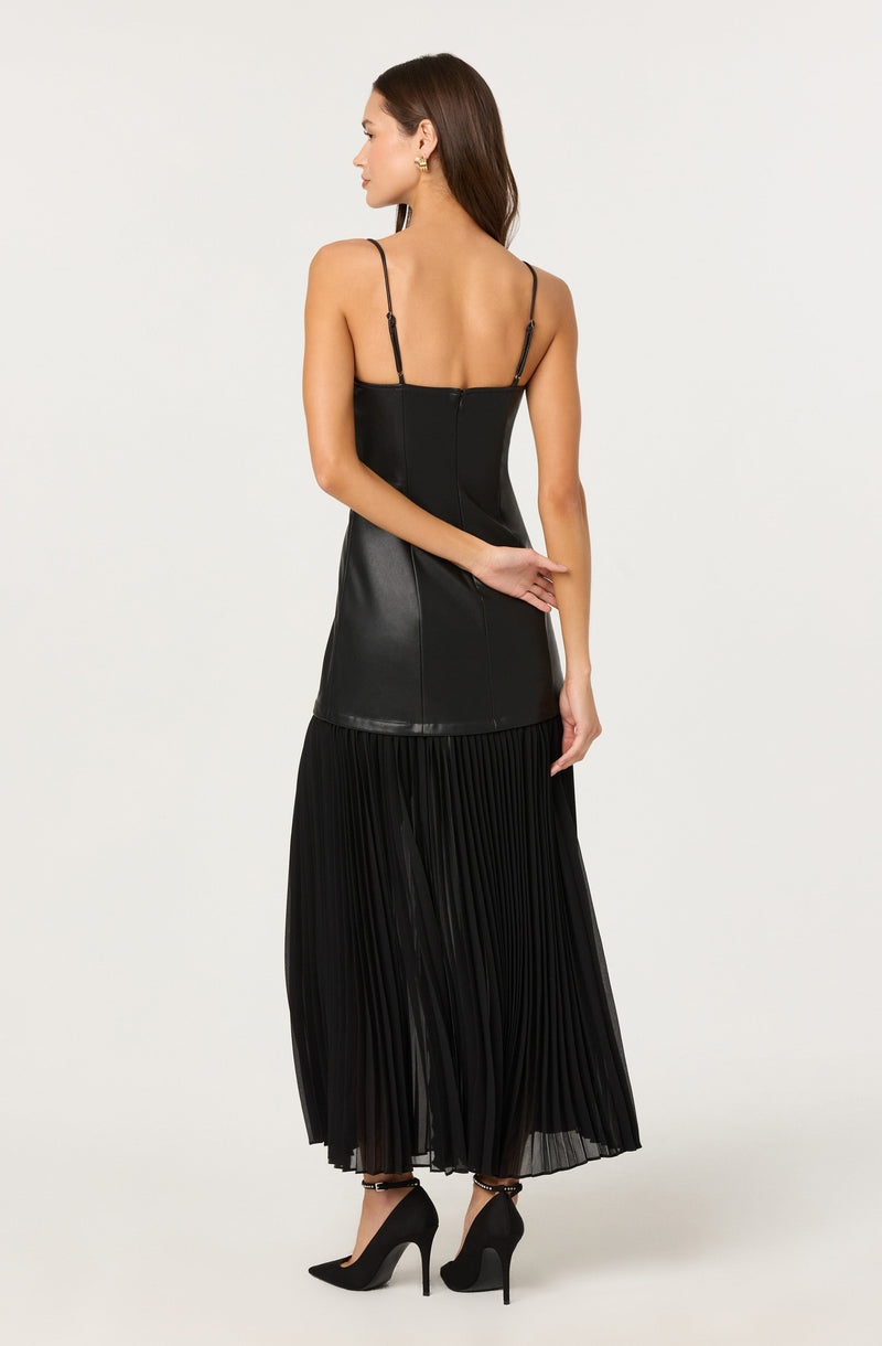 A woman is wearing a black sleeveless dress with thin spaghetti straps. The top part of the dress appears to be made of a fitted, leather-like material that extends to the hips. The lower part of the dress features a pleated, semi-sheer fabric that reaches the floor.