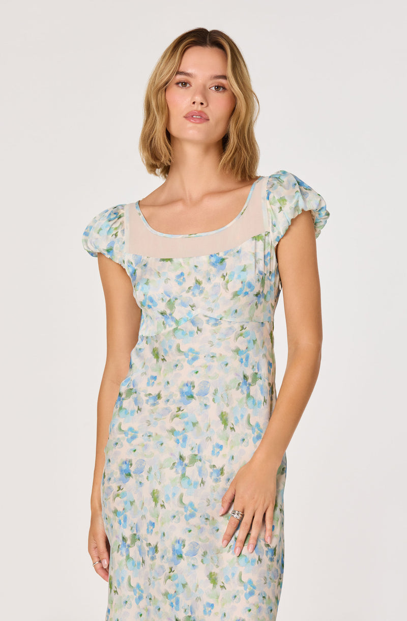 Woman wearing a pastel floral dress with puff sleeves and sheer neckline details.