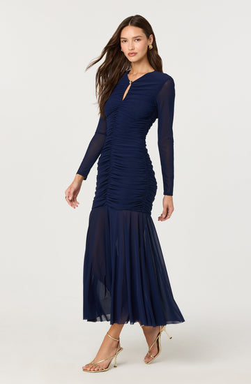 A woman is wearing a long, elegant navy blue dress with a fitted ruched bodice and long sheer sleeves. The dress features a small keyhole opening at the neckline with a metallic clasp and a sheer, flowing skirt that extends to the floor. She is also wearing light-colored, strappy high-heeled sandals.