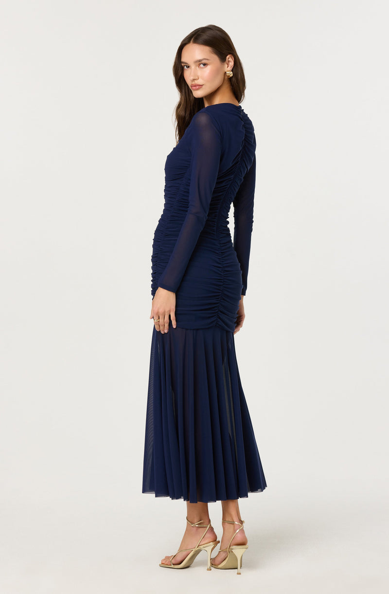 A woman is wearing a long, dark blue dress with a fitted ruched bodice and long sheer sleeves. The skirt features pleated fabric that extends to a midi length. She is accessorized with gold earrings and beige high-heeled sandals.