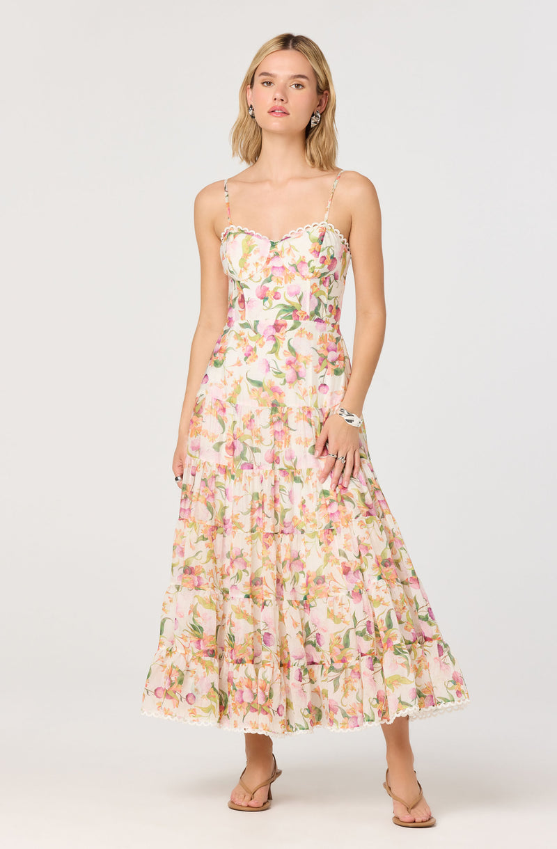 A woman is wearing a sleeveless, ankle-length dress with a floral print featuring pink, yellow, and green colors. The dress has thin spaghetti straps, a fitted bodice with a ruffled neckline, and a tiered skirt with a slightly ruffled hem. She is also wearing open-toed, nude-colored sandals, silver hoop earrings, a bracelet, and rings.