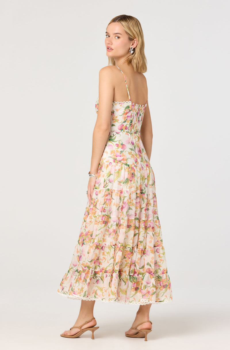 A woman is wearing a sleeveless, ankle-length dress with thin spaghetti straps. The dress features a light, floral pattern in shades of pink, green, and cream, with a smocked bodice and a tiered, ruffled skirt. She is also wearing nude-colored high-heeled sandals and large, decorative earrings.