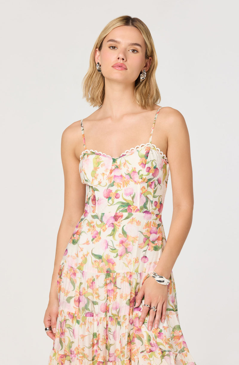A woman is wearing a sleeveless dress with spaghetti straps, featuring a colorful floral pattern in shades of pink, green, and orange. The dress has a ruffled detail at the bust with a scalloped edge along the neckline, and a tiered, flowing skirt. She has blonde, shoulder-length hair styled in loose waves, and she is accessorized with large, statement earrings, a silver bracelet, and rings on her fingers.