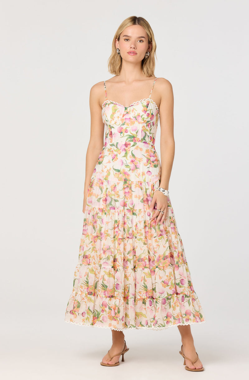 A woman is wearing a sleeveless, ankle-length dress with thin spaghetti straps. The dress features a floral pattern in shades of pink, green, and white, with a ruffled bodice detail and a tiered skirt. She is also wearing beige high-heeled sandals and has accessories including earrings and a bracelet.
