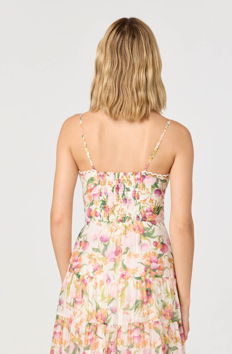 A woman is wearing a sleeveless, spaghetti strap dress with a floral pattern featuring pink, peach, and green colors on a light background. The dress has shirred fabric at the bodice and a tiered skirt with ruffled layers.