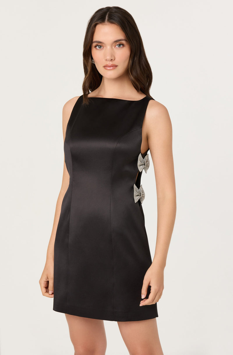 A woman is wearing a sleeveless black dress with a boat neckline. The dress features two decorative bow accents on the side near the waist. The fabric has a smooth, slightly shiny appearance.