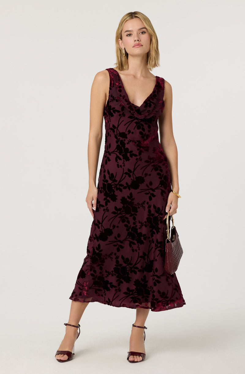A woman is wearing a sleeveless, burgundy velvet dress with a black floral pattern. The dress has a cowl neckline and a fitted waist, flowing down to a slightly flared hem that reaches her ankles. She is accessorizing with gold hoop earrings, a gold bracelet, and is holding a small matching burgundy handbag. She is also wearing strappy high-heeled sandals.