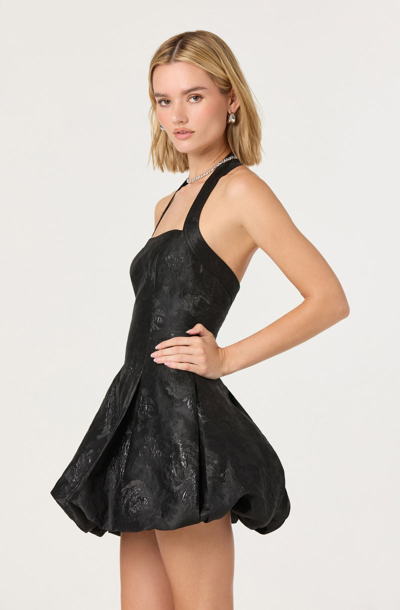 A young woman is wearing a sleeveless black dress with wide straps. The dress has a fitted bodice that extends into a voluminous, bubble-style skirt with a textured, slightly shiny pattern. She accessorizes with silver earrings and a choker necklace.