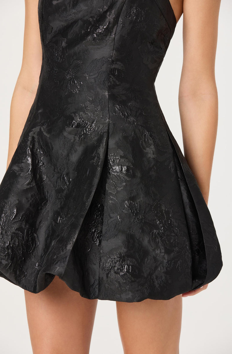 A woman is wearing a sleeveless black dress made from a shiny, textured fabric with a floral pattern. The dress has a fitted bodice and a full, structured skirt with pleats, creating a voluminous silhouette.