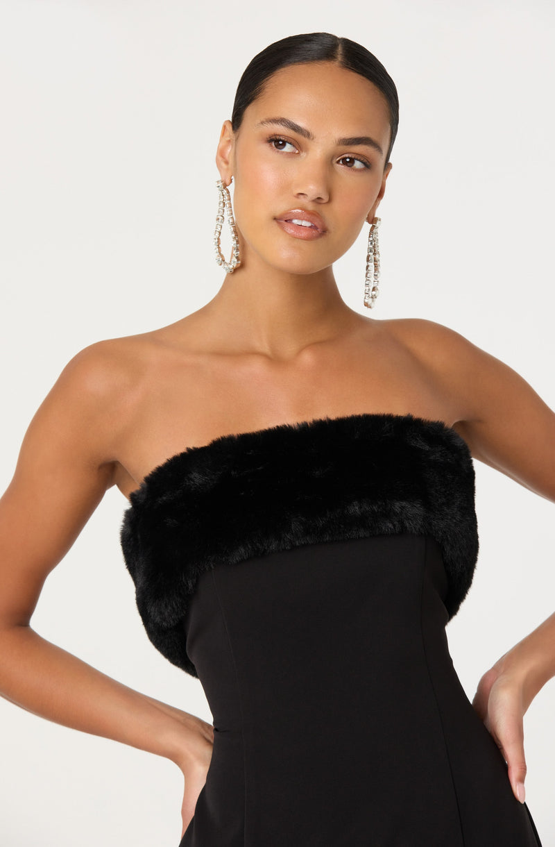 A woman is wearing a strapless black dress with a faux fur trim at the top. She has her hair pulled back and is accessorized with large, dangling earrings that feature multiple strands of sparkling stones.