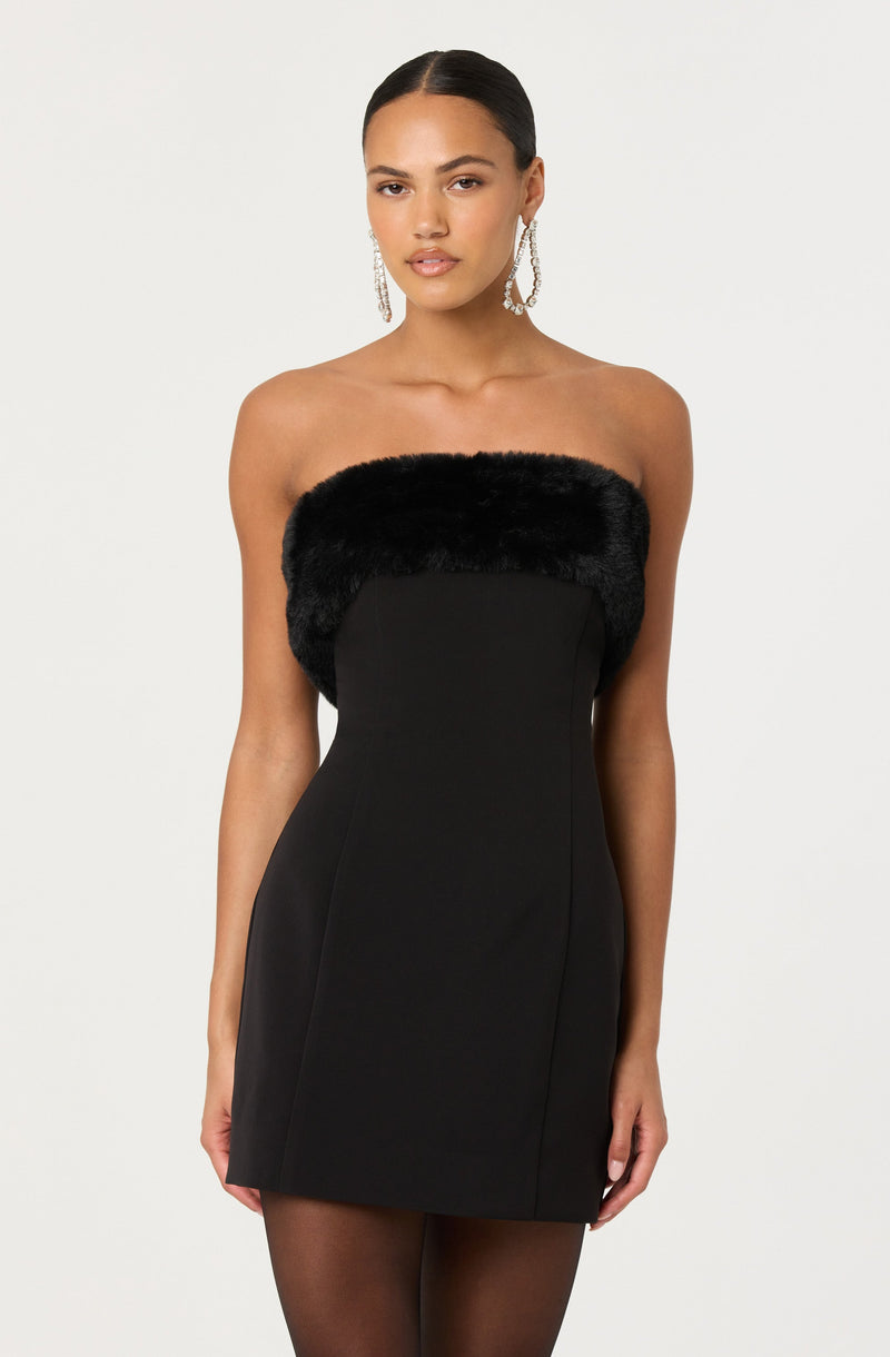 A woman is wearing a strapless black dress with a black faux fur trim around the top. The dress is fitted and short, ending above the knees. She is also accessorized with long, dangling earrings.