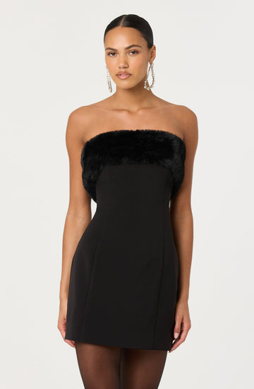 A woman is wearing a strapless black dress with a black faux fur trim around the top. The dress is fitted and short, ending above the knees. She is also accessorized with long, dangling earrings.