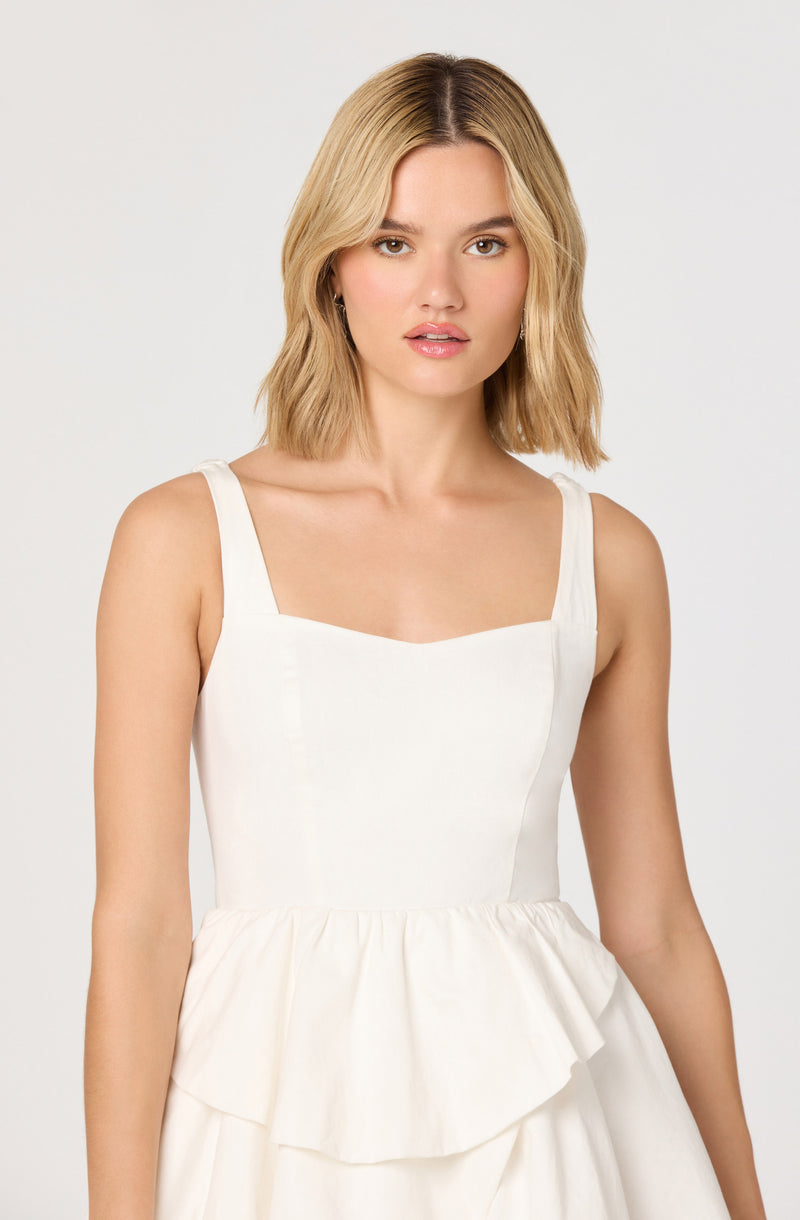 A woman is wearing a sleeveless, white dress with wide shoulder straps and a square neckline. The dress has a fitted bodice and a gathered, ruffled skirt at the waist.