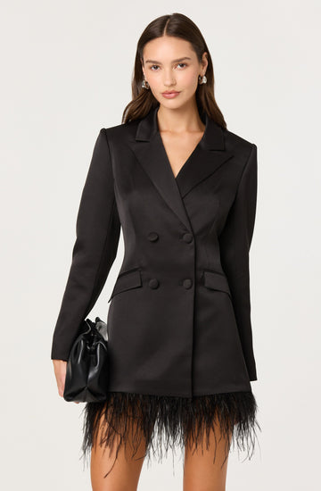 A woman is wearing a black, double-breasted blazer with peak lapels and long sleeves. The blazer features button details and flap pockets at the waist. She pairs it with a feathered black skirt that extends to just above her knees. She is also holding a small black handbag in her right hand.