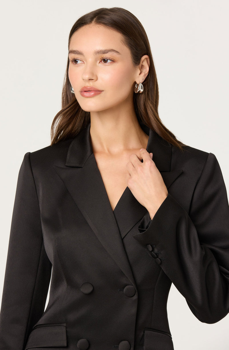 A woman wearing a black, fitted, double-breasted blazer with wide lapels. The blazer has a smooth satin-like finish, three buttons visible on the front, and slightly padded shoulders.
