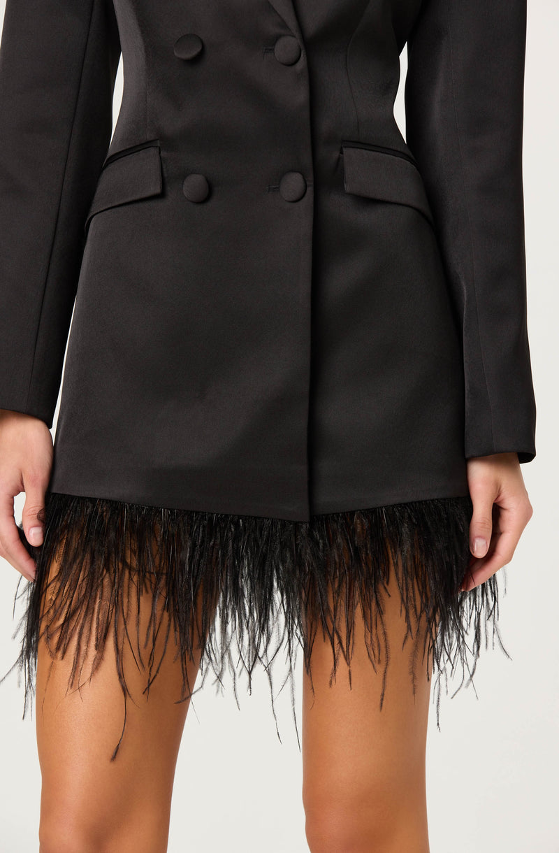 A person is wearing a black tailored blazer with large buttons down the front and flap pockets on each side. The blazer has a fitted style with structured shoulders. They are also wearing a short skirt with a feathered hem that is black at the top and gradually transitions to a lighter, tan color.