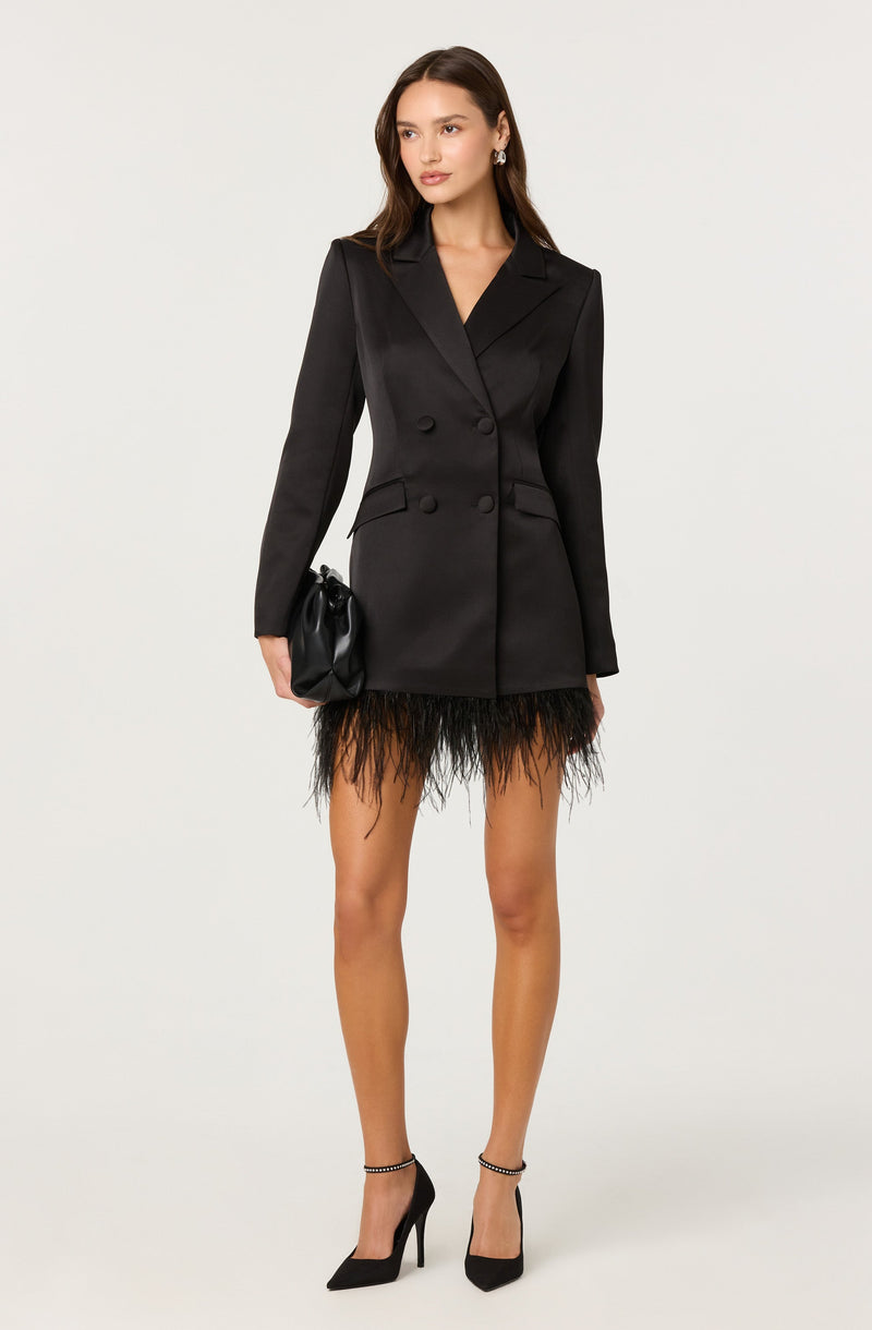 A woman is wearing a black, double-breasted blazer with wide lapels and structured shoulders, paired with a short, black feathered skirt that has a fringed appearance. She is holding a small black bag in her right hand and is wearing black high heels with ankle straps.