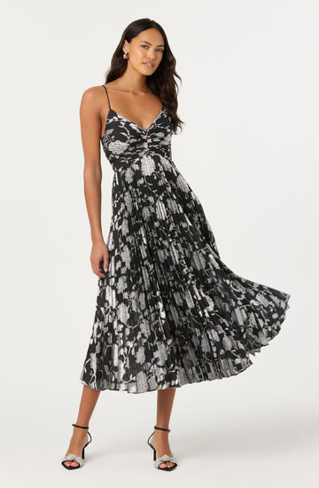 A woman is wearing a sleeveless, black dress with thin spaghetti straps. The dress features a V-neckline and a fitted bodice, with a flowy, pleated skirt that reaches mid-calf. The fabric has a black and silver abstract animal print pattern. She pairs the dress with black and white high-heeled sandals.