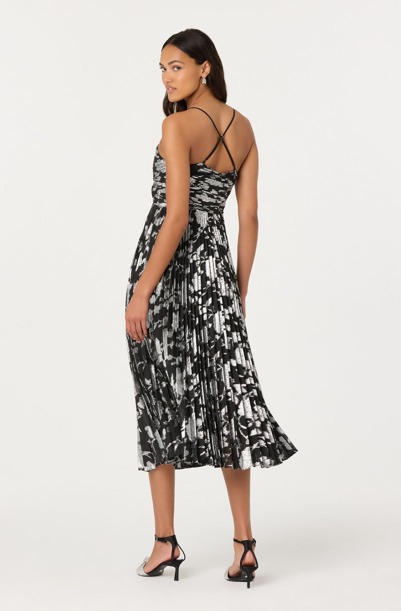 A woman is wearing a sleeveless, form-fitting black and white patterned dress with thin, crisscross spaghetti straps on the back. The dress has a smocked bodice and a pleated midi-length skirt with an abstract, geometric design. She is also wearing black high-heeled sandals with ankle straps.