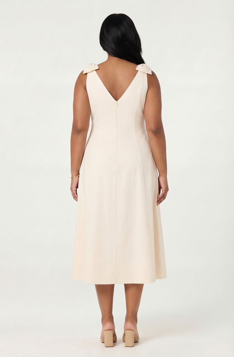 Back view of a woman wearing a sleeveless cream-colored midi dress with bows on shoulders and beige high heels.
