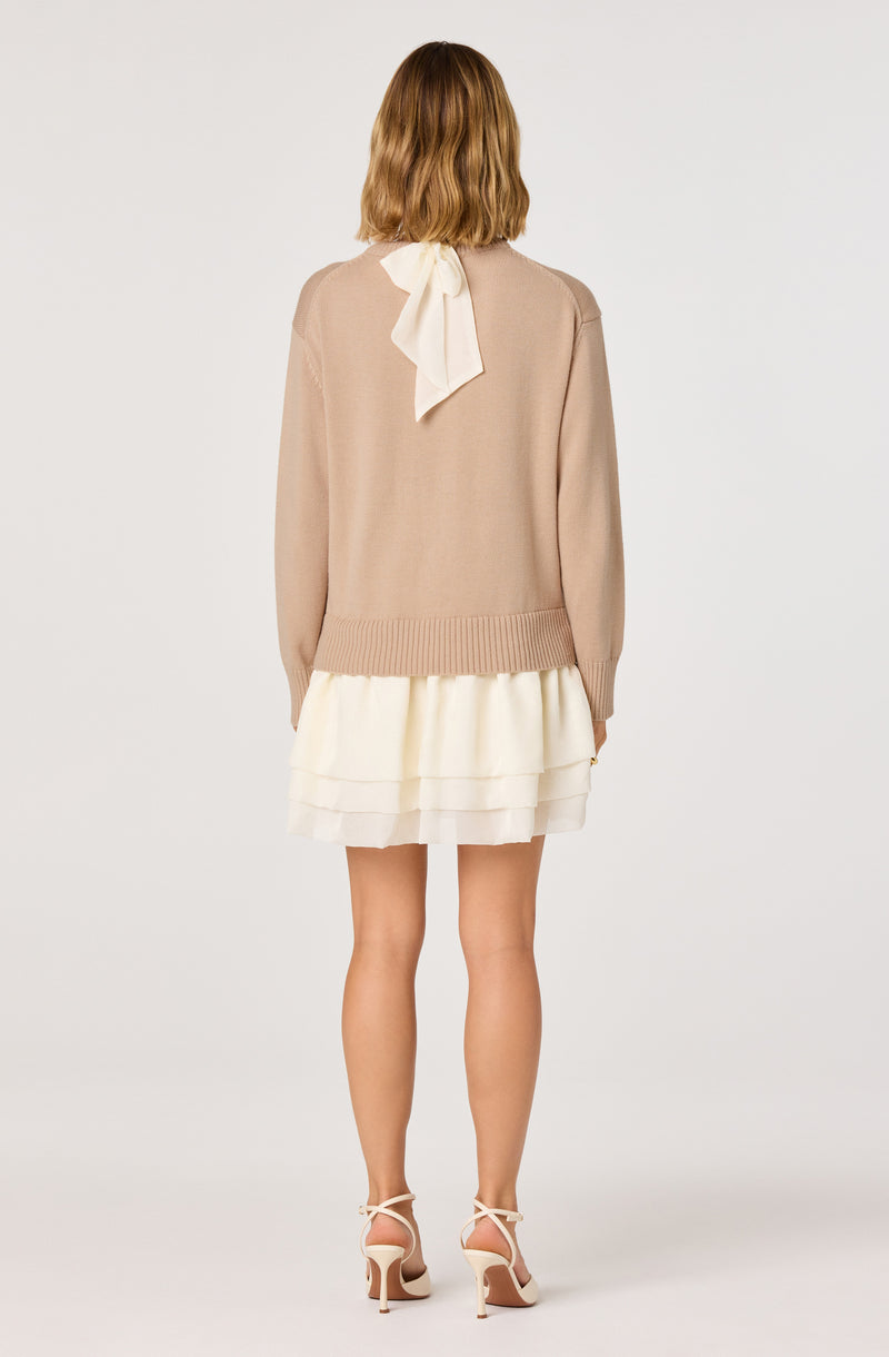 Woman wearing a beige sweater with a white bow at the back and a white layered skirt.