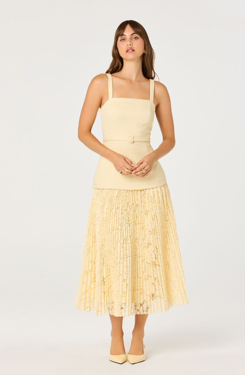 Woman wearing a beige sleeveless top with shoulder straps and a matching belt, paired with a yellow pleated lace skirt and beige heels.