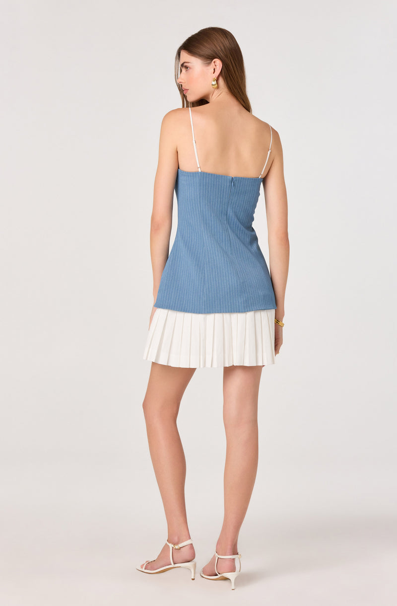 Woman in a blue sleeveless top with white spaghetti straps and a cream pleated skirt.