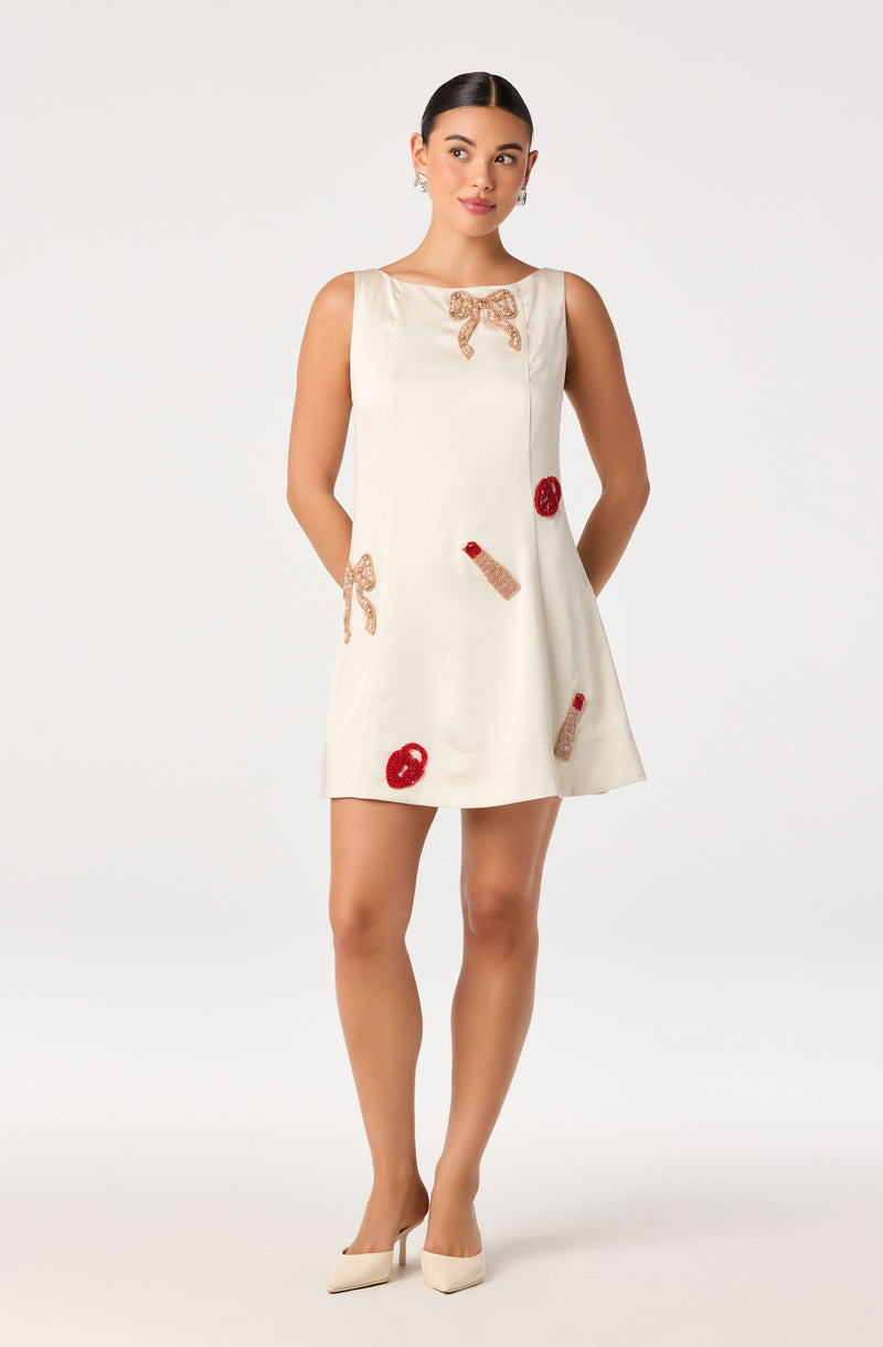 Cream sleeveless dress with embroidered bows, lips, and lipstick motifs, and beige heels.