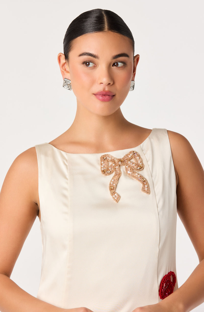 Woman wearing a sleeveless cream dress with a pearl-encrusted bow and red embellished detail.