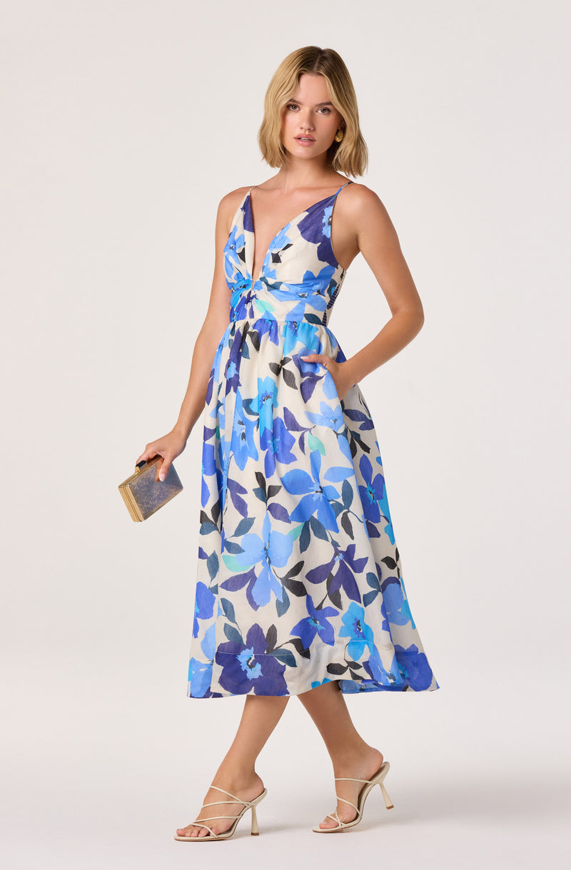 Woman wearing a sleeveless, V-neck, floral dress in shades of blue, with a fitted waist and flowy skirt.