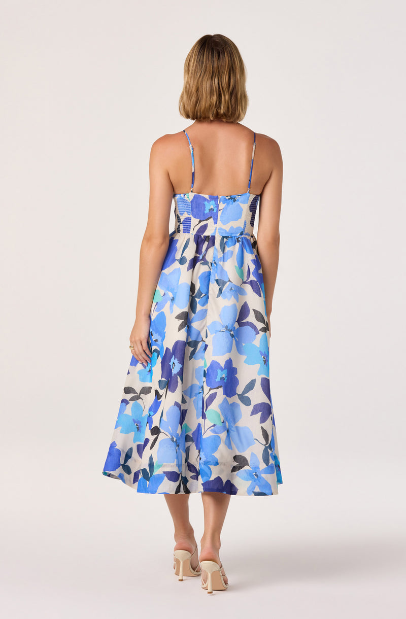 Woman wearing a sleeveless dress with thin straps and a blue, purple, black, and beige floral pattern.