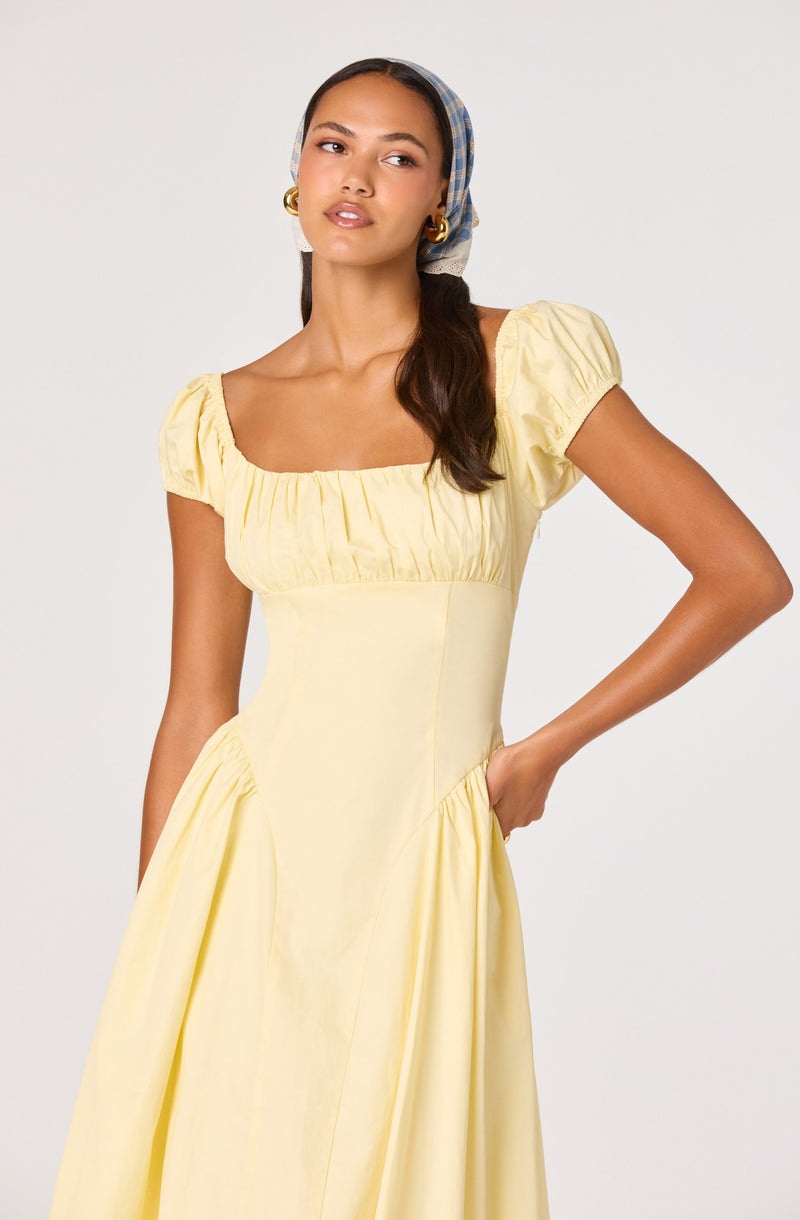 Woman in a light yellow dress with puff sleeves and a fitted bodice. She wears a blue checked headscarf and gold hoop earrings.