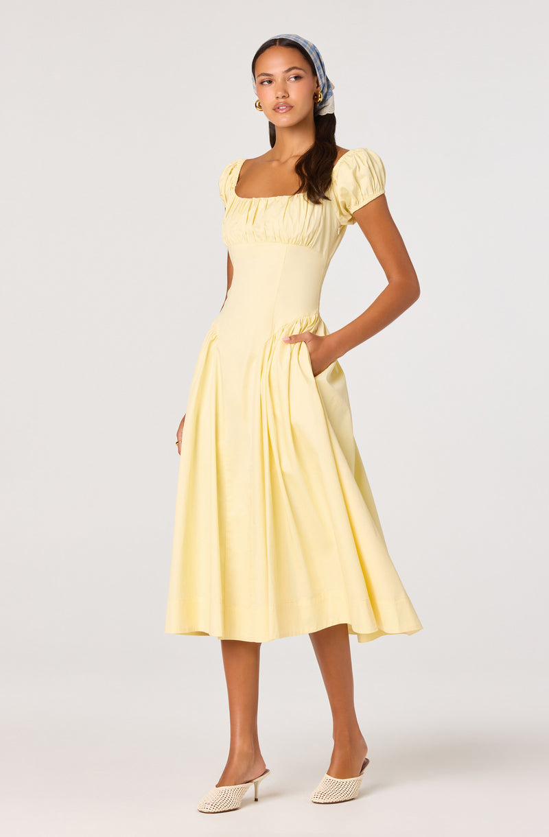 Woman in a yellow midi dress with puff short sleeves, square neckline, and side pockets, paired with woven heels and a patterned headscarf.