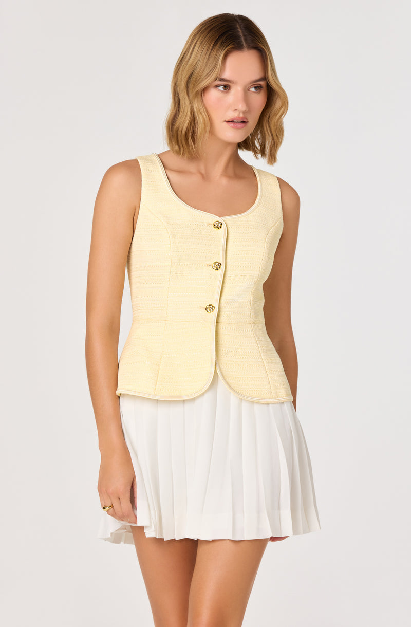Woman in sleeveless yellow textured jacket with gold buttons and a white pleated skirt.