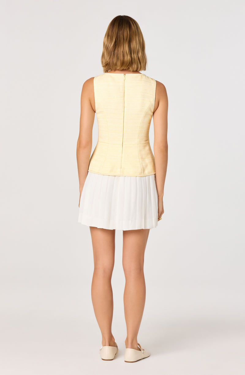 Woman wearing a sleeveless, yellow textured top and a white pleated skirt, with beige shoes.