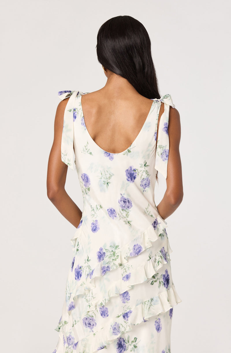 Woman in a white floral dress with purple flowers and ruffled layers, shoulder ties.