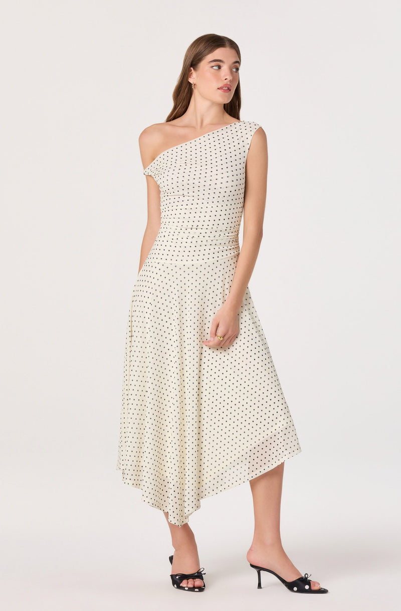 Woman in a beige polka dot dress with one shoulder sleeve and black open-toe heels.