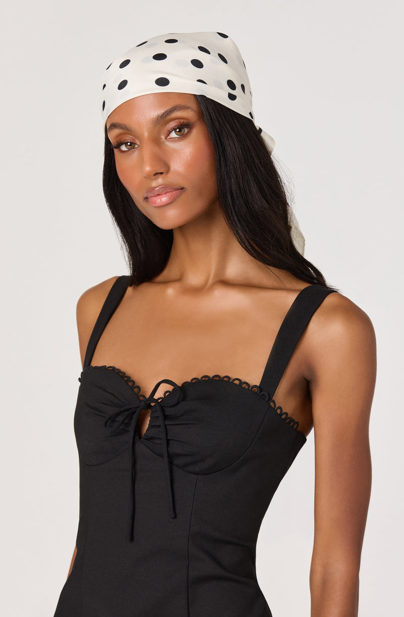 Woman in a black dress with bow detail and scalloped trim, wearing a white polka dot headscarf.