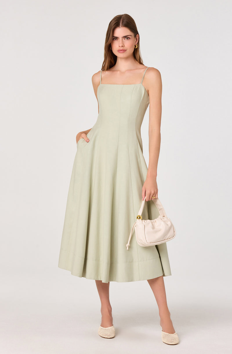 Woman wearing a light green, sleeveless, spaghetti strap midi dress with pleats, holding a white purse.