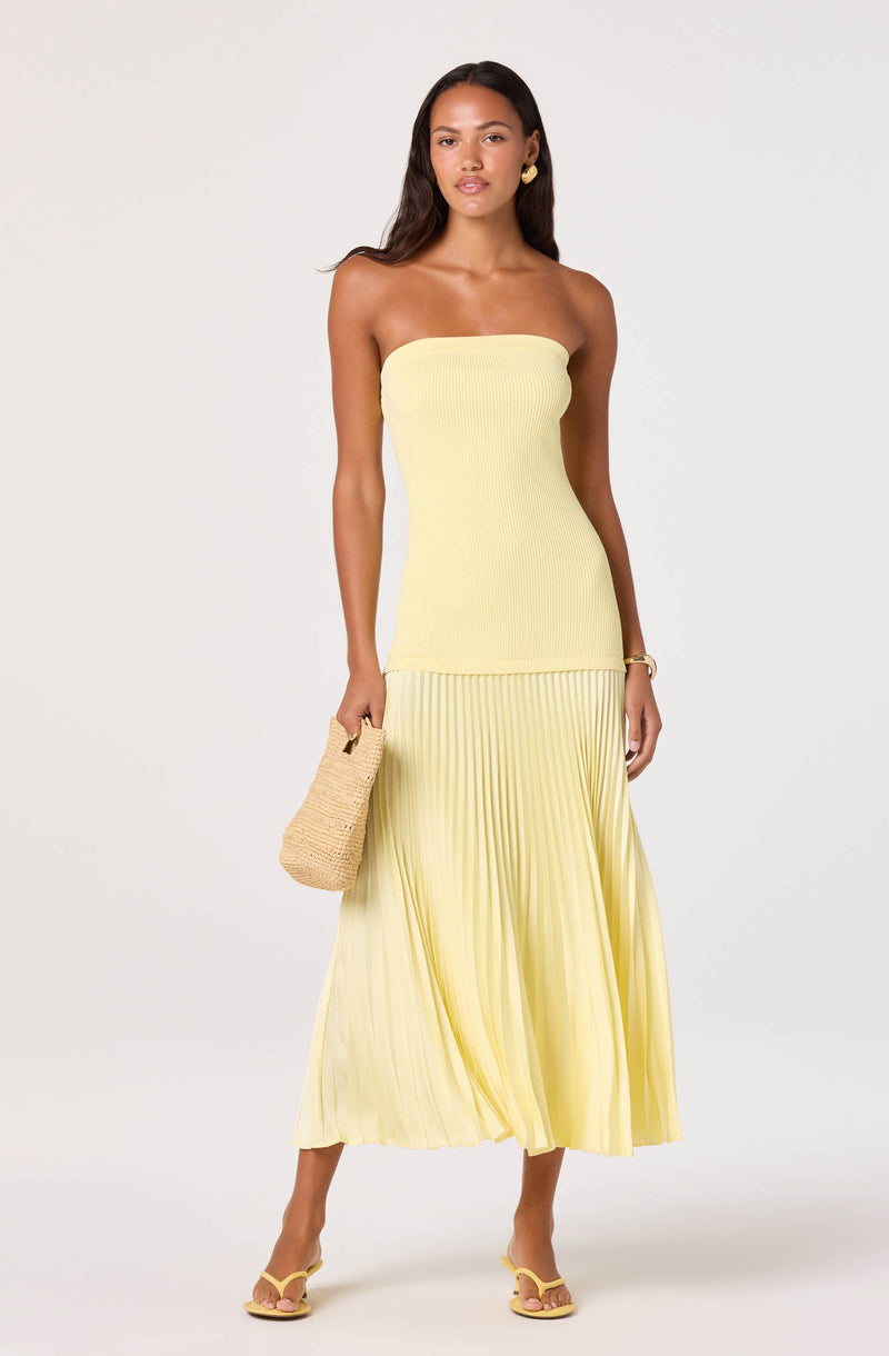 Woman wearing a strapless, yellow, ribbed top with a matching pleated, flowy midi skirt, holding a straw handbag.