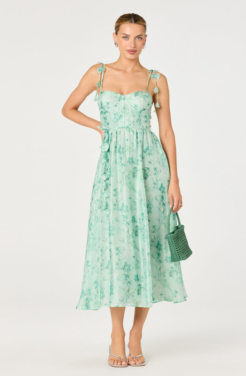 Woman wearing a sleeveless, ankle-length, light green dress with floral pattern and tied shoulder straps.