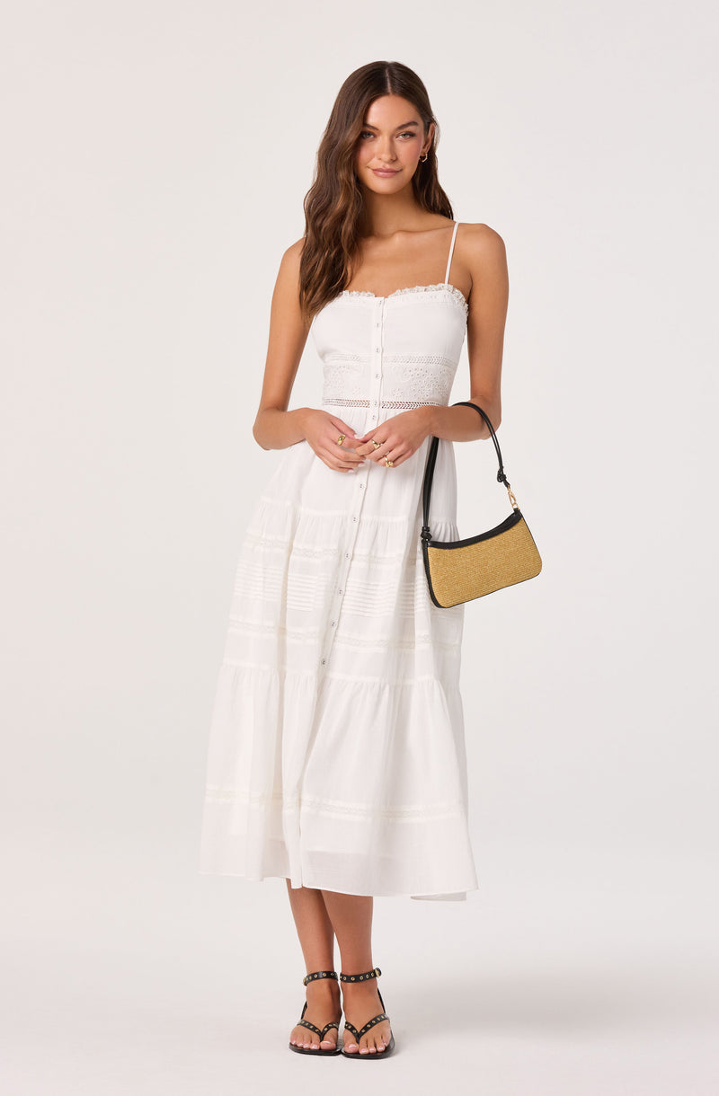 Woman in a white, sleeveless, tiered maxi dress with spaghetti straps, holding a beige and black handbag.